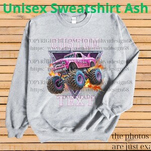 Op de afbeelding: Asgrijze unisex sweatshirt met een roze monstertruck ontwerp met blauwe en roze wielen. Het ontwerp bevat de tekst "CUSTOM TEXT" en "Unisex Sweatshirt Ash". De sweatshirt wordt getoond op een houten achtergrond.