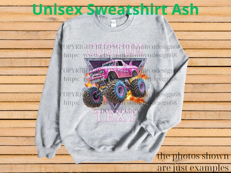Op de afbeelding: Asgrijze unisex sweatshirt met een roze monstertruck ontwerp met blauwe en roze wielen. Het ontwerp bevat de tekst "CUSTOM TEXT" en "Unisex Sweatshirt Ash". De sweatshirt wordt getoond op een houten achtergrond.