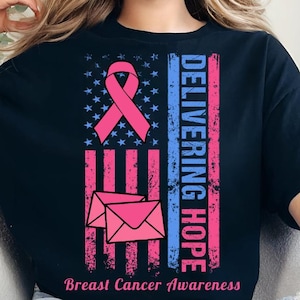 May include: Black t-shirt with a pink ribbon, envelope graphic, and text "DELIVERING HOPE" and "Breast Cancer Awareness". The design incorporates a vertical American flag motif with blue and pink stripes and a star field.