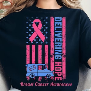 May include: Black t-shirt with a pink ribbon and the text "DELIVERING HOPE" in blue and pink. The design includes a blue mail truck and the words "Breast Cancer Awareness". The shirt has a distressed American flag design.