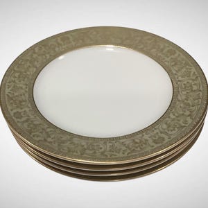May include: A stack of four white ceramic plates with a gold rim and a decorative olive green border. The border features a repeating floral pattern. The plates are stacked on top of each other.