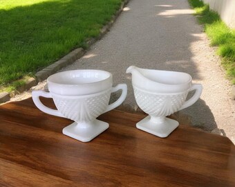 May include: A white milk glass creamer and sugar bowl set. The sugar bowl has two handles and the creamer has a spout. Both pieces have a diamond pattern and sit on square bases. The set is on a dark wood surface.