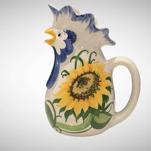 May include: A ceramic pitcher with a rooster design. The pitcher is white with blue and yellow accents. A large sunflower is painted on the front, with green vines and leaves. The handle is curved and white.