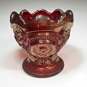 May include: A ruby red, iridescent glass bowl with a scalloped rim and a pedestal base. The bowl features embossed floral and geometric patterns. The glass has a shimmering, metallic finish, reflecting gold and orange hues.
