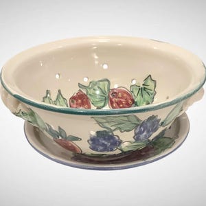 May include: Cream-colored ceramic colander with a matching saucer base. The colander features hand-painted illustrations of strawberries, blueberries, and green leaves. The rim and base are outlined in teal.