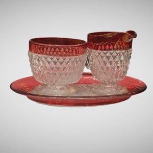 May include: A vintage sugar and creamer set with a matching tray. The set features clear glass with a diamond pattern and a red rim. The tray and rims are a vibrant red. The creamer has a handle and spout.