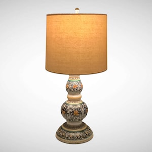 May include: A decorative table lamp with a beige lampshade and a ceramic base. The base features a detailed floral design in shades of black, orange, and green. The lamp has a round lampshade and a clear glass finial.