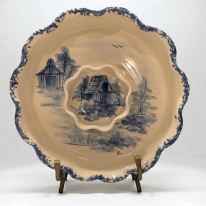 May include: A cream-colored ceramic serving dish with a blue barnyard scene and scalloped edges. The dish has a smaller, matching bowl in the center. The rim and edges of the dish are trimmed with a blue pattern. The dish is displayed on a wooden stand.