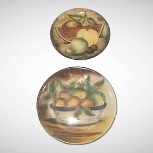 May include: Two decorative ceramic plates with hand-painted fruit designs. One plate features a watermelon slice, mango, and other fruits. The second plate depicts a bowl of oranges and other fruits. The plates have a brown and yellow color scheme.