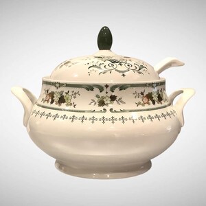 May include: A white ceramic soup tureen with a matching lid and two side handles. The tureen features a dark green finial on the lid and a decorative band of green floral patterns and intricate scrollwork around the body and the lid edge.