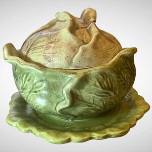 May include: A ceramic cabbage-shaped serving dish with a lid, resting on a matching scalloped plate. The dish and lid are primarily green with brown accents, and the design mimics the texture of cabbage leaves.