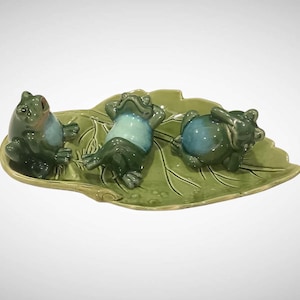 May include: A ceramic decorative tray shaped like a green leaf featuring three small frog figurines. The frogs have glossy green and light blue bodies, with one sitting upright and two others lying down in relaxed positions on the leaf surface.
