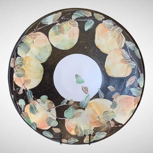 May include: A round, decorative plate with a dark brown background and a white center. The plate is adorned with a painted design of yellow and orange fruit with green leaves and brown stems. The plate is displayed on a black metal stand.