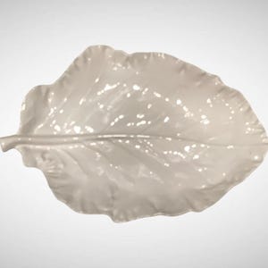 May include: A glossy, off-white ceramic leaf-shaped dish. The dish has a detailed vein pattern and a gently curved, ruffled edge. The neutral color and organic shape make it suitable for serving or as a decorative accent.