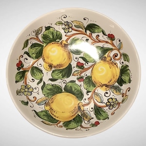 May include: A round, off-white ceramic bowl with a hand-painted design. The bowl features three large yellow fruit with green leaves and red berries, all connected by brown vines. The rim is slightly curved, and the overall design is detailed and colorful.