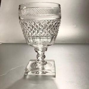 May include: Clear crystal goblet with a diamond-cut pattern on the bowl and a decorative band near the rim. The goblet has a faceted stem and a square base. The glass reflects light, creating a sparkling effect.