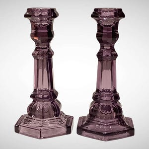 May include: Two matching purple glass candlesticks. Each candlestick has a square base, a fluted column, and a decorative top. The glass has a faceted design, creating a play of light and shadow. These decorative candlesticks are ideal for adding a touch of elegance to any room.