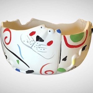 May include: White ceramic bowl with a cat face design. The bowl features a whimsical design with red, green, blue, and black abstract shapes and swirls. The cat's face has red spots and a black nose and mouth.