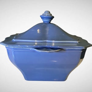 May include: A blue ceramic lidded serving dish. The dish has a square shape with a curved, scalloped edge and a decorative finial on the lid. The color is a solid, muted blue.