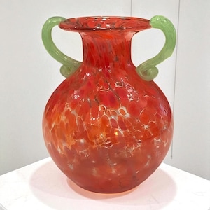 May include: A vibrant red glass vase with a bulbous body and two green handles. The vase has a speckled pattern with white and orange accents. The handles are a bright green color. The vase is a decorative item.