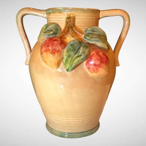 May include: A ceramic vase with two handles, decorated with relief-molded fruit and leaves. The vase is primarily beige with green and peach accents. The handles and rim are a light peach color. The vase is a decorative home decor item.