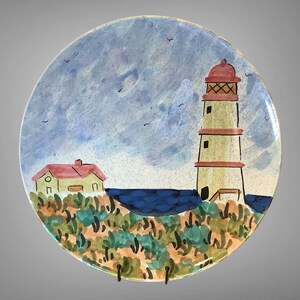 May include: A round ceramic plate featuring a hand-painted coastal scene. The design includes a lighthouse with a red top, a small house, and a blue ocean. The plate is decorated with a sky blue background and colorful foliage.