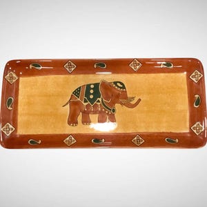May include: Rectangular ceramic serving tray with a brown border and a tan center. The tray features an elephant design in the center, with a brown body, green and black accents, and a decorative border with floral and paisley patterns.