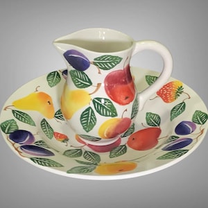 May include: A white ceramic pitcher and matching dish set. The pitcher has a curved handle and spout. Both pieces are decorated with colorful illustrations of fruits, including plums, pears, and strawberries, with green leaves.