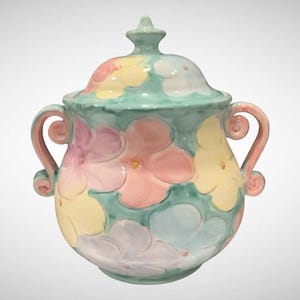 May include: A ceramic jar with a lid, painted with pastel flowers in pink, yellow, and blue on a teal background. The jar has two pink handles and a small finial on the lid. The overall design is whimsical and decorative.