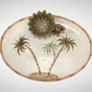 May include: An oval serving platter with a bamboo-style rim. The ivory-colored surface features painted palm trees and a small monkey figurine. A green leaf-shaped bowl sits at the top.