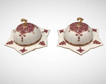 May include: Two white ceramic butter dishes with lids, each adorned with a burgundy and gold floral design. The lids have a gold handle. The dishes sit on star-shaped plates with matching burgundy floral patterns.