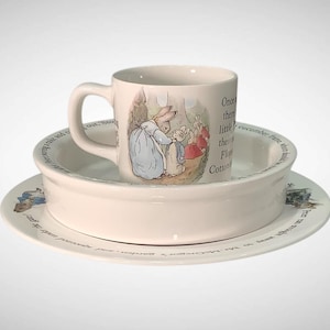 May include: A three-piece ceramic dish set featuring illustrations of rabbits in a garden scene. The set includes a mug, a bowl, and a plate, all with a cream-colored background and a classic children's book theme.