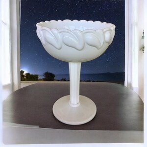May include: A white, stemmed serving bowl with a scalloped rim and leaf-like designs. The bowl sits on a round base and a fluted stem. The background shows a night sky with stars.