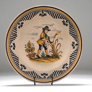 May include: A decorative ceramic plate featuring a hand-painted scene of a person harvesting wheat. The plate has a cream-colored base with a black and yellow border. The central image depicts a figure in a hat and colorful clothing.