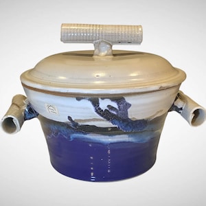 May include: A ceramic casserole dish with a lid and two handles. The dish is primarily white with blue and brown accents. The base is a deep blue color. The lid has a handle. The dish is handmade.