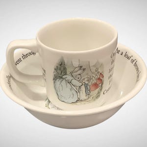 May include: A white ceramic mug and bowl set. The mug features a scene with rabbits and text. The bowl and mug are off-white. The mug is sitting inside the bowl. The set is likely for children.