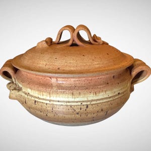 May include: A ceramic lidded casserole dish with a speckled, earthy color palette. The lid features a decorative looped handle, and the dish has curved handles on either side. The colors range from light tan to reddish-brown.