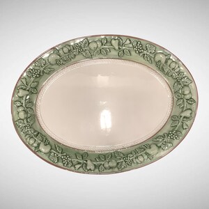 May include: An oval ceramic serving platter with a smooth, cream-colored center and a wide, sage green border. The border features a raised, embossed relief pattern of various fruits, including pears and grapes, surrounded by detailed leaves.