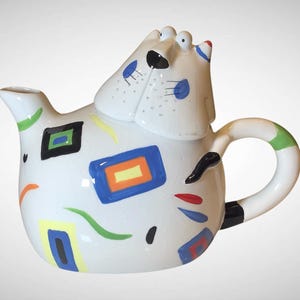 May include: A whimsical white ceramic teapot with a cat-like design. The teapot features a rounded body, a curved handle, and a spout. It is decorated with colorful geometric shapes in blue, orange, green, and black. The lid is shaped like the cat's head.