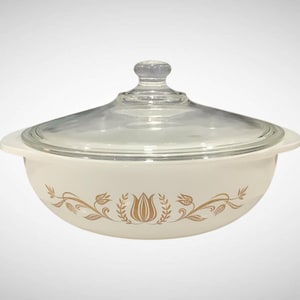 May include: Cream-colored, covered casserole dish with a clear glass lid. The dish features a gold floral design around the base. The lid has a clear glass knob. A vintage kitchenware item.