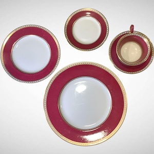 May include: A set of fine china with a white center and a deep red border. The border is accented with a gold trim. The set includes a teacup and saucer, two plates, and a smaller plate.