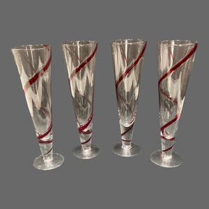 May include: A set of four clear glass flutes, each featuring a tapered body and a circular base. The glass is decorated with a distinct red spiral pattern that winds around the exterior, creating a continuous line from the base to the rim.