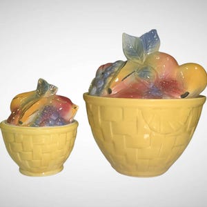 May include: Two yellow ceramic fruit bowls with lids. The lids are shaped like a colorful arrangement of fruit, including grapes, peaches, and pears. The bowls have a basket weave design.