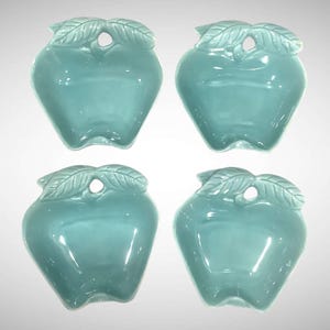 May include: A set of four matching ceramic dishes shaped like apples in a glossy light teal finish. Each dish features a molded stem and leaf detail at the top with a small circular cutout, creating a uniform appearance across all four pieces.