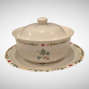 May include: A cream-colored ceramic serving bowl with a matching lid and saucer. The set features a decorative border of green leaves and small red hearts, with a central illustration of a birdhouse, a small tree, and a flying bird.