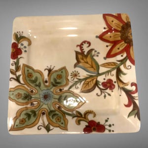 May include: A square ceramic plate with a cream-colored base featuring a painted floral pattern. The design includes a large green and yellow flower in the lower left, a large red and yellow flower in the upper right, and red accents.