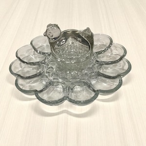May include: A clear glass serving dish with a central bowl and surrounding petal-shaped compartments. The dish has a decorative handle and is designed for serving snacks or appetizers. The glass has a slight blue tint.