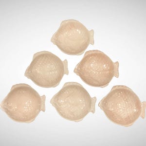 May include: Seven small, fish-shaped dishes in a pale pink hue are arranged on a white surface. The dishes have textured scales and fins, and are likely decorative or for serving small portions.