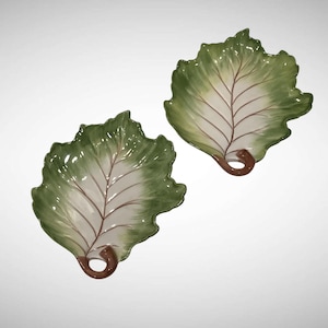 May include: Two leaf-shaped ceramic serving dishes. The dishes are painted with a green and white color scheme, with brown veins and stems. The edges of the leaves are scalloped. The dishes are designed to resemble lettuce leaves.