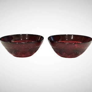 May include: Two matching deep red glass bowls featuring a pressed geometric pattern on the exterior. The bowls have a circular rim and a tapered base, with a textured design consisting of intersecting lines and fan-shaped motifs in the glass.